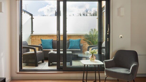 A room with a sliding glass door leading to an outdoor seating area with wicker furniture and a small table with a vase. - Hammersmith Mews by Viridian Apartments (London)