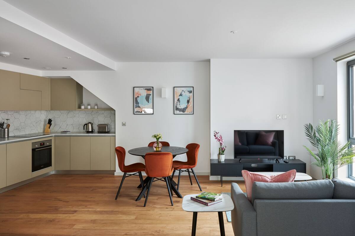 A modern kitchen with a dining area, a living room with a sofa and TV, and a large window.