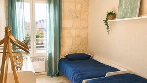 A room with two beds, a wooden chair, a hanging light, a stone wall, a window with curtains, and a plant on a shelf.