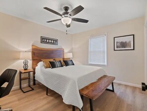 Een slaapkamer met een bed, een plafondventilator, een nachtkastje, een stoel, een raam met jaloezieën en een ingelijst beeld aan de muur.