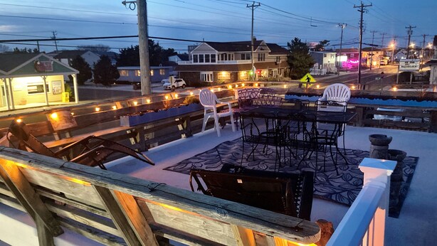 Terraza en la azotea con mesas al aire libre, un edificio pequeño al fondo y una calle con vehículos y letreros iluminados.
