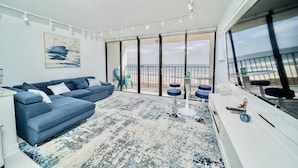 Un salon moderne avec un canapé bleu, un tapis à motifs et un balcon offrant une vue sur la plage.