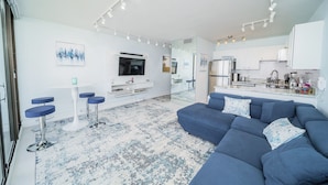 Un salon moderne avec un canapé bleu, un téléviseur et un coin cuisine doté de meubles blancs.