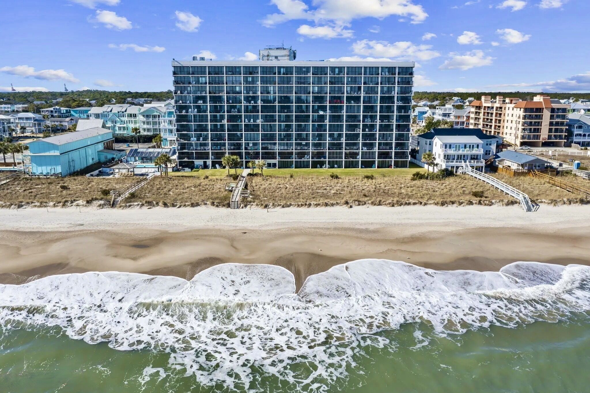 A beachfront hotel with a modern glass facade, a sandy beach in the foreground, and a clear sky.
