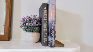 A white shelf with three books titled 'Patricia Cornwell: Predator', 'A novel by the Painter of Light', and 'Kane'. A small potted plant with purple flowers is also visible. - The Cottage on 5th (Eastman)