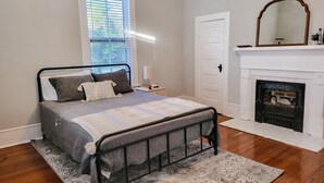 Ein Schlafzimmer mit einem Bett, einem Nachttisch, einem Spiegel, einem Kamin und einem Fenster mit Jalousien.