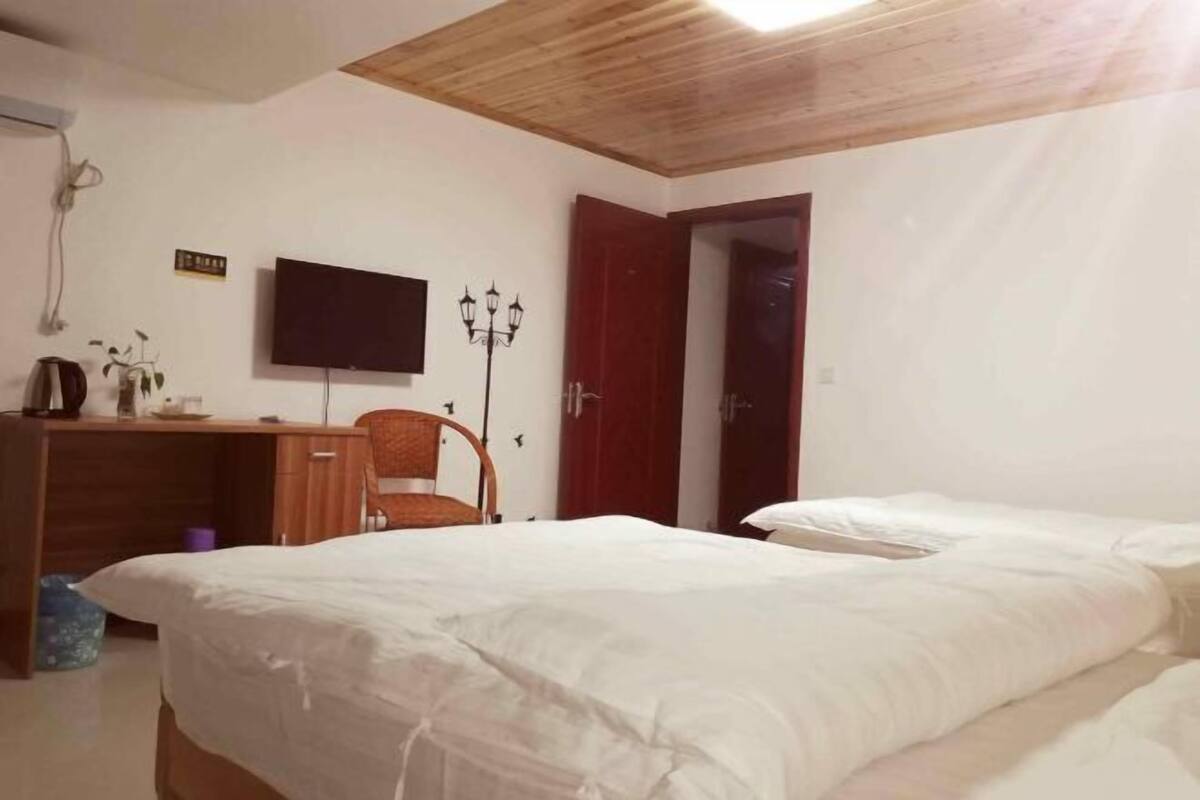 Un dormitorio con techo de madera, un televisor montado en la pared, un escritorio con silla y una cama con ropa de cama blanca.