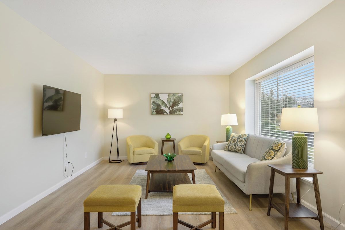 A living room with a flat-screen TV, two armchairs, a sofa, a coffee table, and a window with blinds.