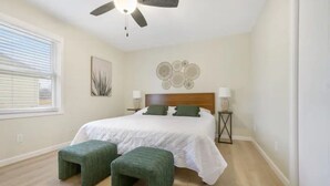 Une chambre à coucher comprenant un lit, deux tables de chevet, un ventilateur de plafond, une fenêtre avec des stores et une œuvre d’art murale.