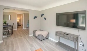 Ein modernes Wohnzimmer mit einem Flachbildfernseher, einem grauen Sofa, einem kleinen Tisch und einem Essbereich im Hintergrund.