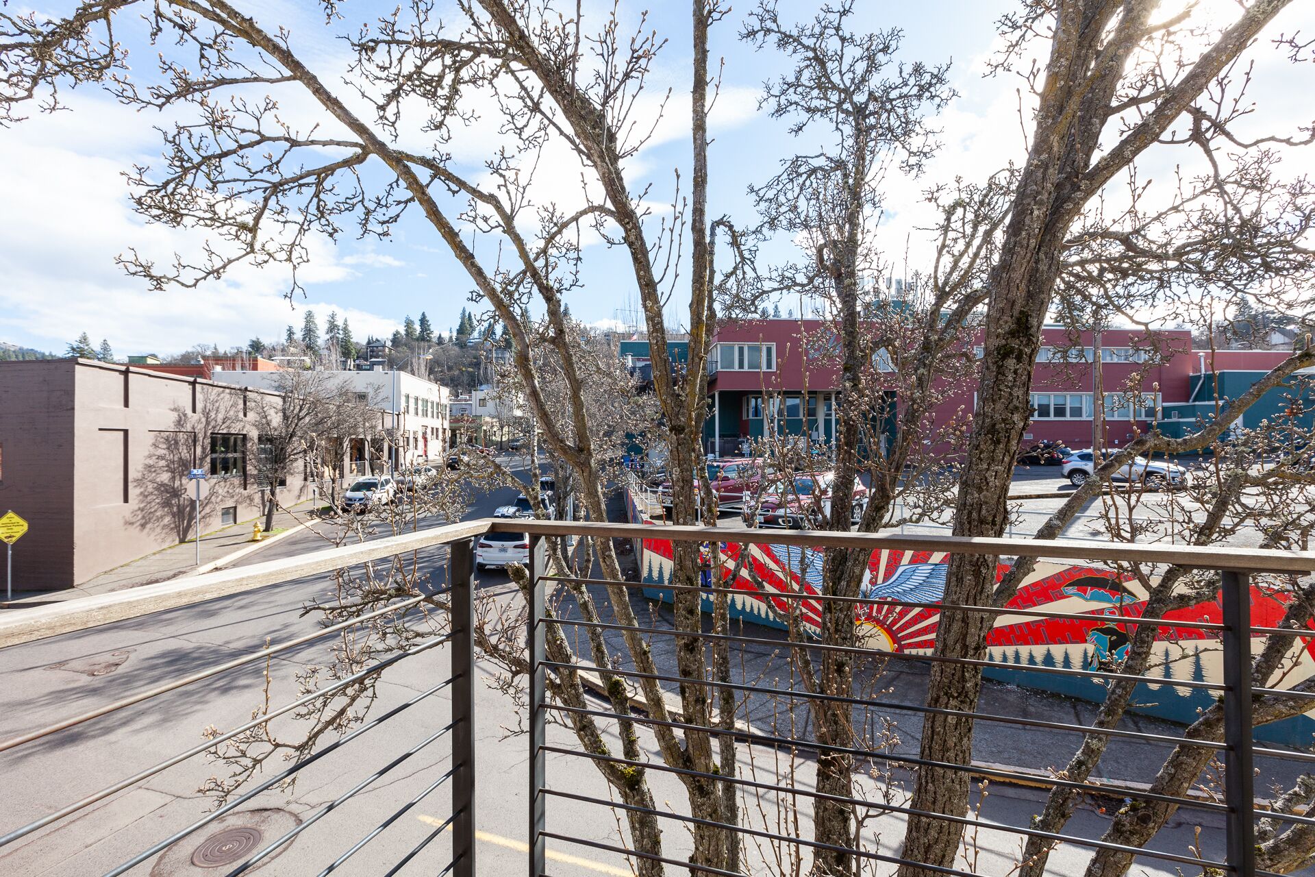 A view from a balcony overlooking a street with a mural, buildings, and parked cars.