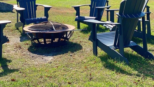Un campamento con sillas Adirondack y un brasero, rodeado de un área verde con árboles en tonos otoñales.