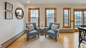 Una habitación con dos sillones azules, una mesita con una lámpara y una mesa de comedor con sillas. Hay ventanas amplias con marcos de madera y vistas al exterior.