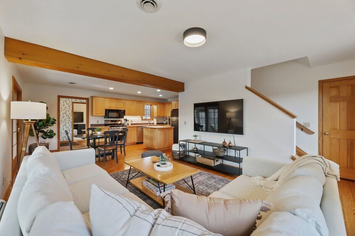 A modern living room with a sofa, a coffee table, and a TV. There is a kitchen in the background with wooden cabinets and a dining area.