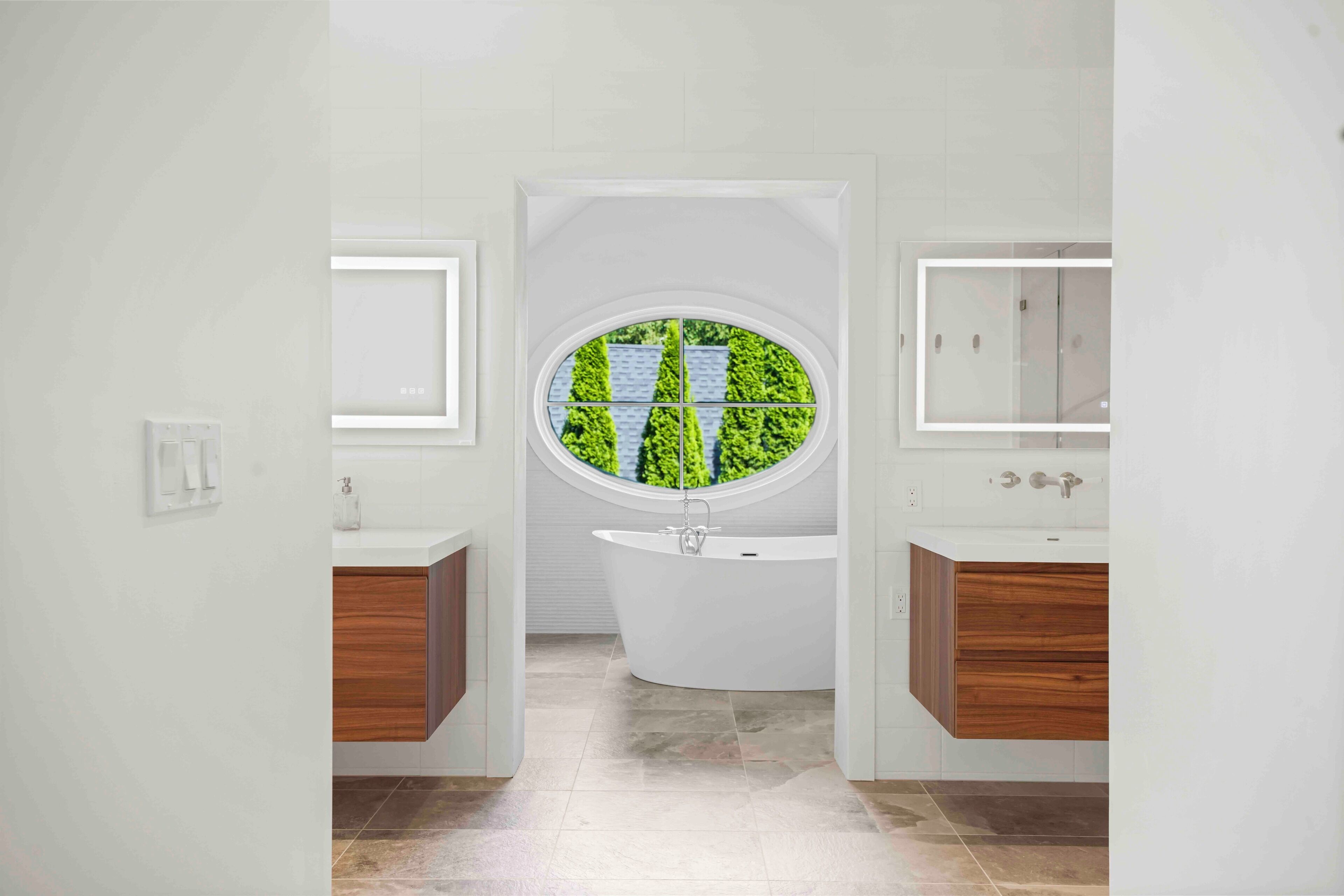 Een moderne badkamer met een vrijstaand bad, twee spiegelkastjes en een groot raam met uitzicht op groene bomen.