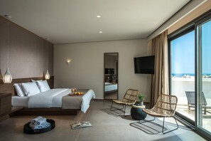 A modern hotel room with a large bed, a seating area with two chairs, a television, and a balcony with a view of the sea. - Metropole Urban Hotel (Heraklion)