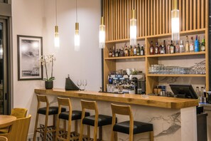 A bar area with wooden bar stools, a counter with a coffee machine, and shelves with bottles and glasses. - Metropole Urban Hotel (Heraklion)