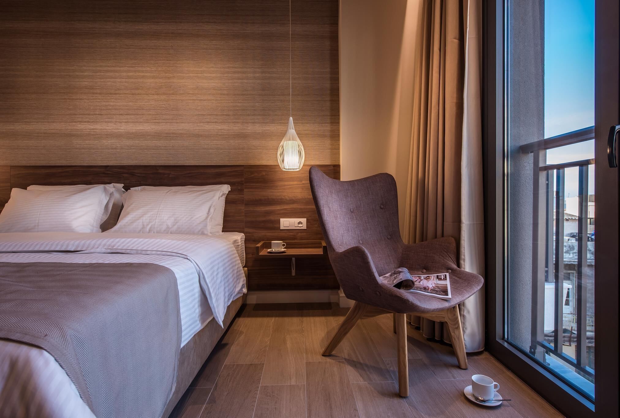 Een hotelkamer met een bed, een stoel, een nachtkastje, een hanglamp en een raam met gordijnen.
