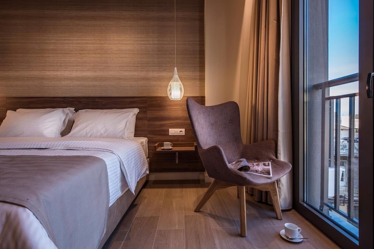 Une chambre d’hôtel avec un lit, une chaise, une table de chevet, une lampe suspendue et une fenêtre avec des rideaux.