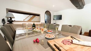 A dining area with a table set for four, including plates of food, glasses of wine, and a bottle of wine. There are two chairs on each side of the table, and a television mounted on the wall above. - Small village house in the heart of Normandy! (Ferrières-la-Verrerie)