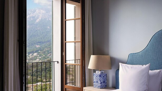 Ein Hotelzimmer mit Bett, Nachttisch mit Lampe und Blick auf eine Berglandschaft durch ein offenes Fenster.