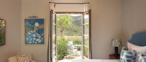 A bedroom with a bed, a chair, a nightstand, a painting, and a view of an outdoor patio.