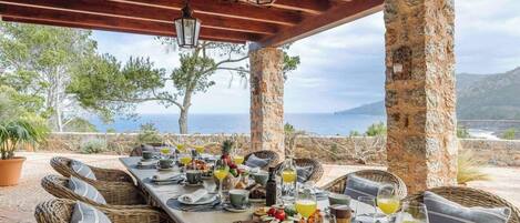 Un espace extérieur aménagé pour les repas, avec des piliers en pierre, des meubles en osier et une vue sur la mer.