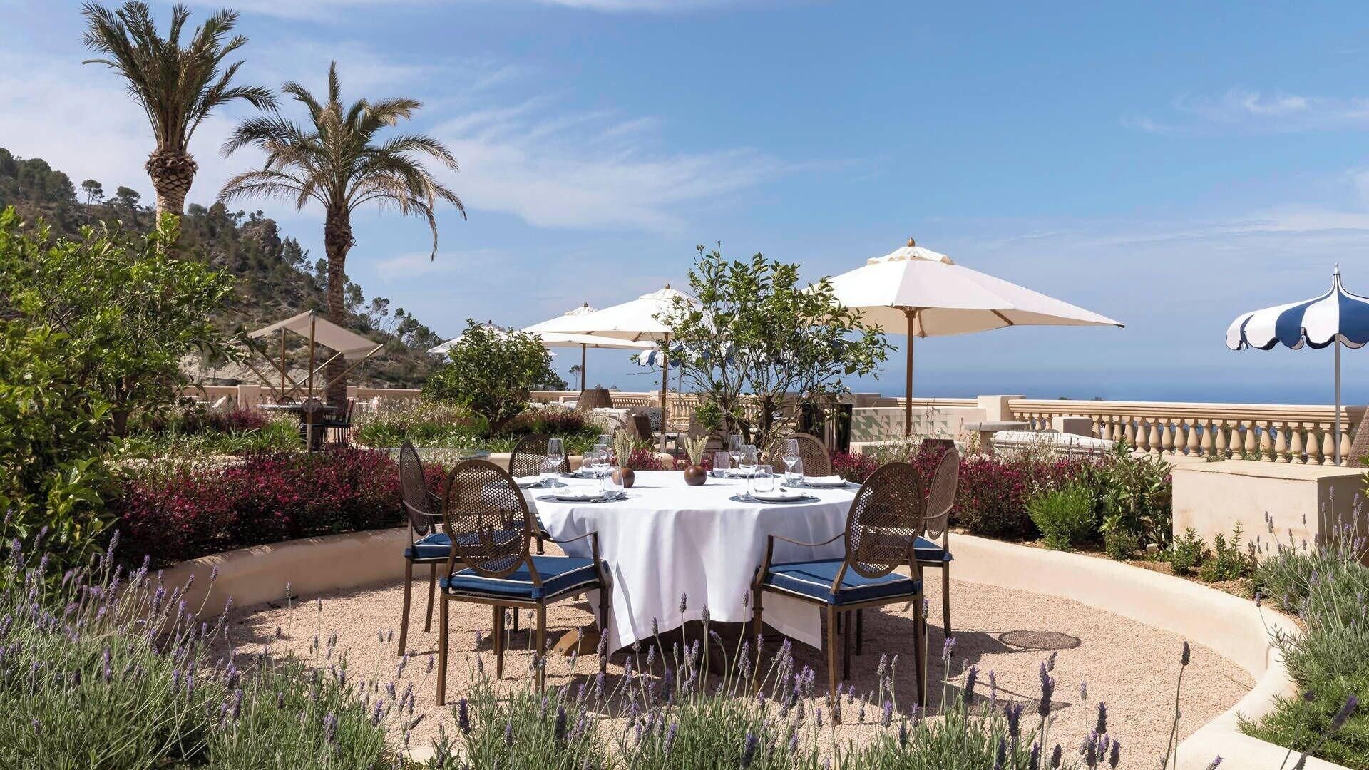 Un espace de restauration en plein air, avec des tables disposées pour deux personnes, entouré de plantes et de palmiers, et offrant une vue panoramique.