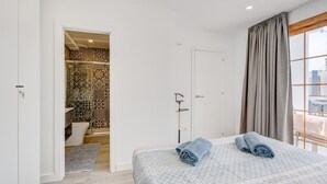 Een slaapkamer met een bed, een badkamer met betegelde wanden, een raam met gordijnen en uitzicht op gebouwen.