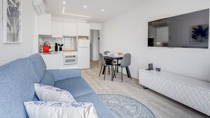 Een moderne woonkamer met een blauwe bank, een eettafel met stoelen, een keuken met witte kasten en een tv aan de muur.