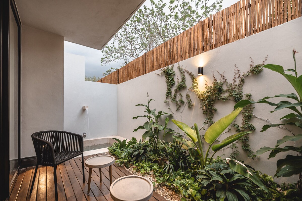 Una terraza de madera con una silla de mimbre negro y dos mesas redondas, rodeada de abundante vegetación y una valla de madera.