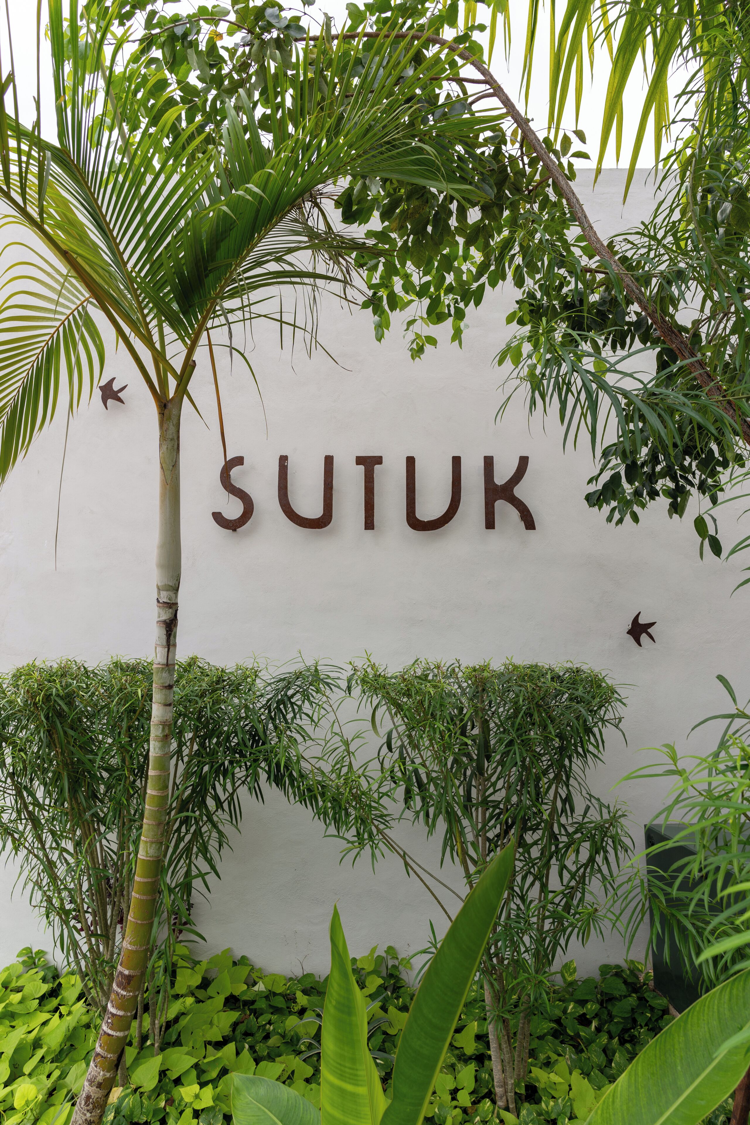 Ein Schild mit der Aufschrift "SUTUK", umgeben von üppigem Grün und Pflanzen.