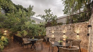 En udendørs patio med trægulv, fletstole og borde, omgivet af frodig vegetation og en stenvæg.