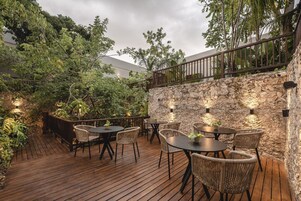 Un patio al aire libre con suelo de madera, sillas y mesas de mimbre, rodeado de abundante vegetación y un muro de piedra.