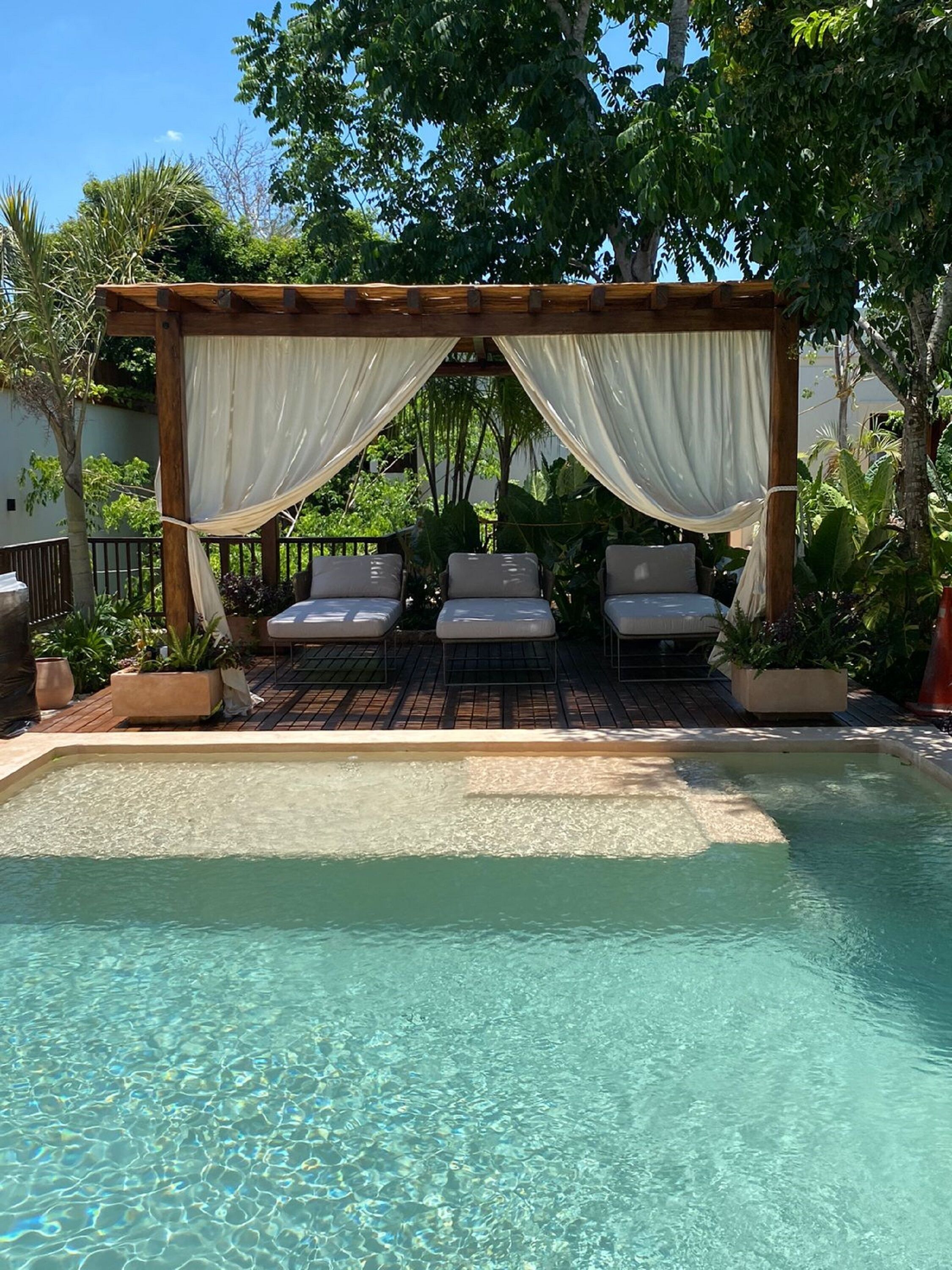 A pool area with a pergola, three lounge chairs, and a clear pool.