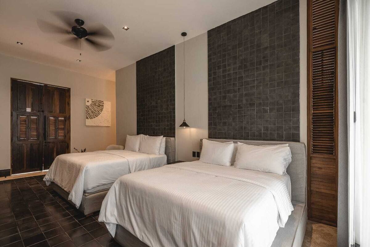 Habitación de hotel con dos camas, ventilador de techo, pared de acento con azulejos oscuros y una puerta de madera.