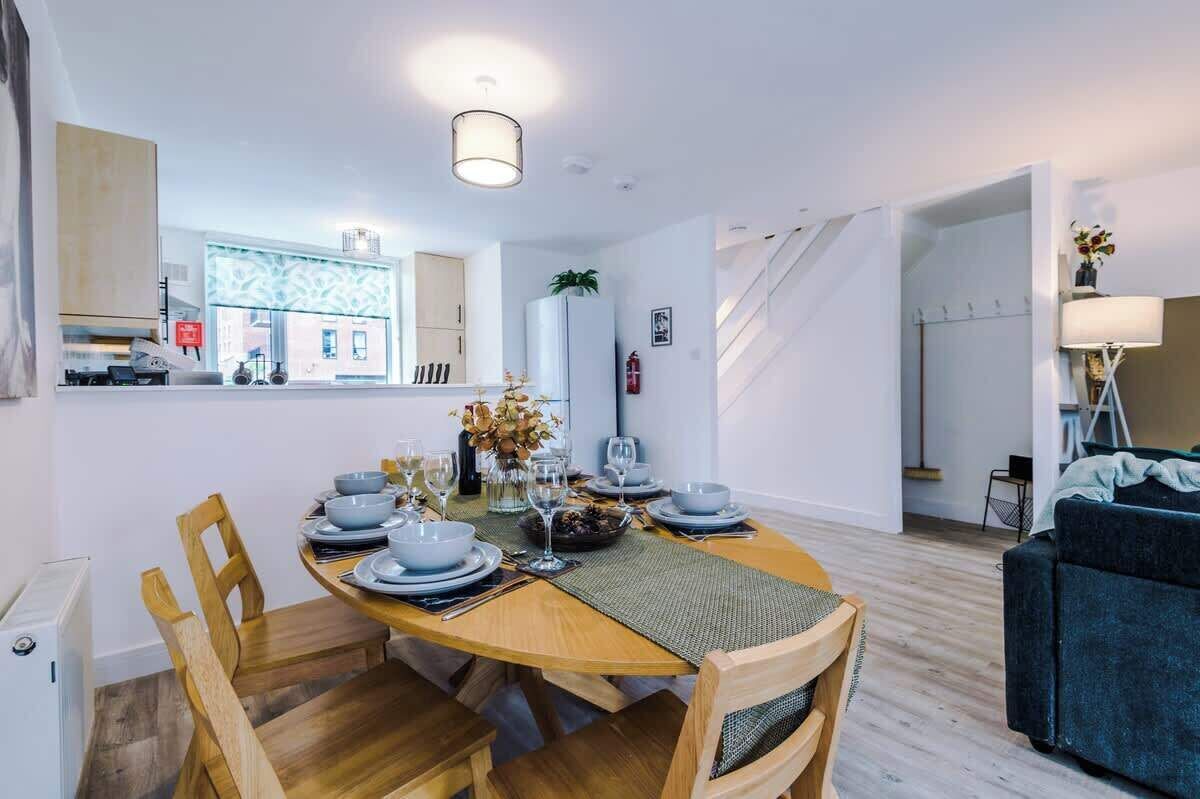A modern dining area with a wooden table set for four, surrounded by wooden chairs. A kitchen with white cabinets and a patterned backsplash is visible in the background.