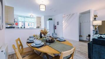 A modern dining area with a wooden table set for four, surrounded by wooden chairs. A kitchen with white cabinets and a patterned backsplash is visible in the background.