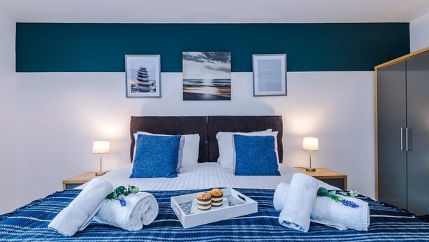 A neatly made bed with blue and white bedding, two pillows, and a tray with muffins. There are two bedside lamps, a dark headboard, and a teal accent wall with framed pictures. - Spacious House, Incredible Location, FREE Parking! (Chester)