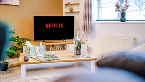A cozy living room with a wooden coffee table, a remote control, a bottle of champagne, and a glass. A TV screen displays the Netflix logo. - Spacious House, Incredible Location, FREE Parking! (Chester)