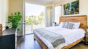 A bedroom with a wooden headboard, a large bed, a balcony with a view of greenery, and a painting on the wall.