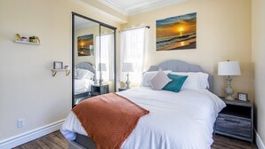 A bedroom with a bed, a nightstand, a mirror, a shelf with decorative items, and two sunset paintings on the wall.