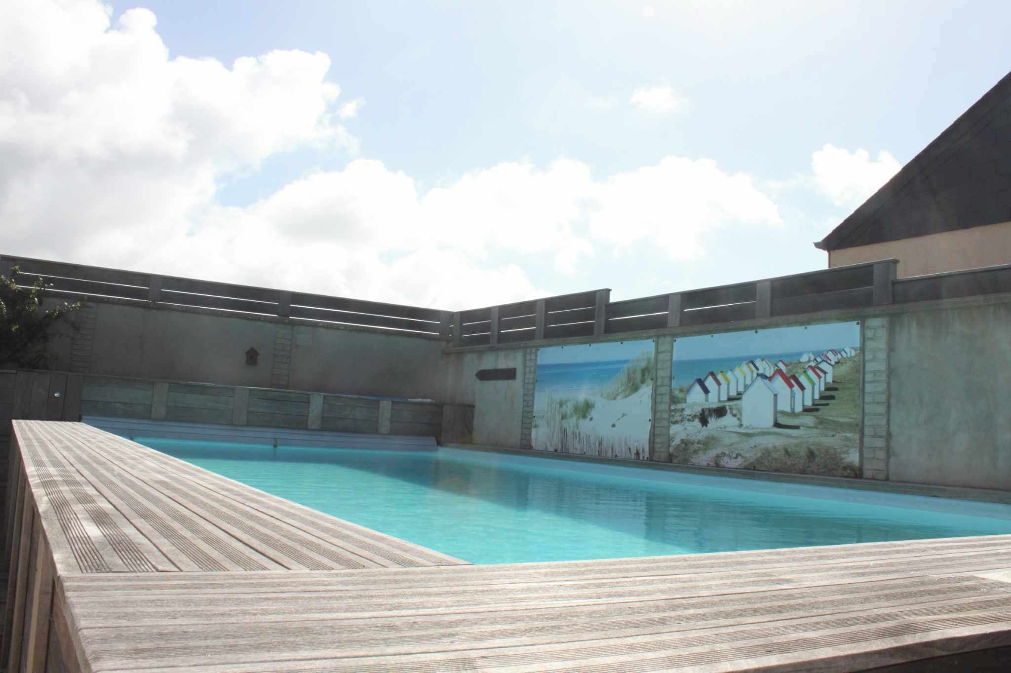 Una piscina con cubierta de madera, un mural de casas de playa y cielo despejado.