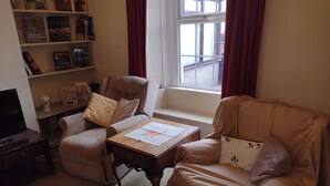 A living room with a sofa, armchair, and a coffee table. There is a bookshelf with books and decorative items. A window with curtains is visible. - Remarkable 3-bed House in Millom (Millom)