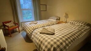 A bedroom with two beds, a window with curtains, a chair, and a bedside table. - Remarkable 3-bed House in Millom (Millom)
