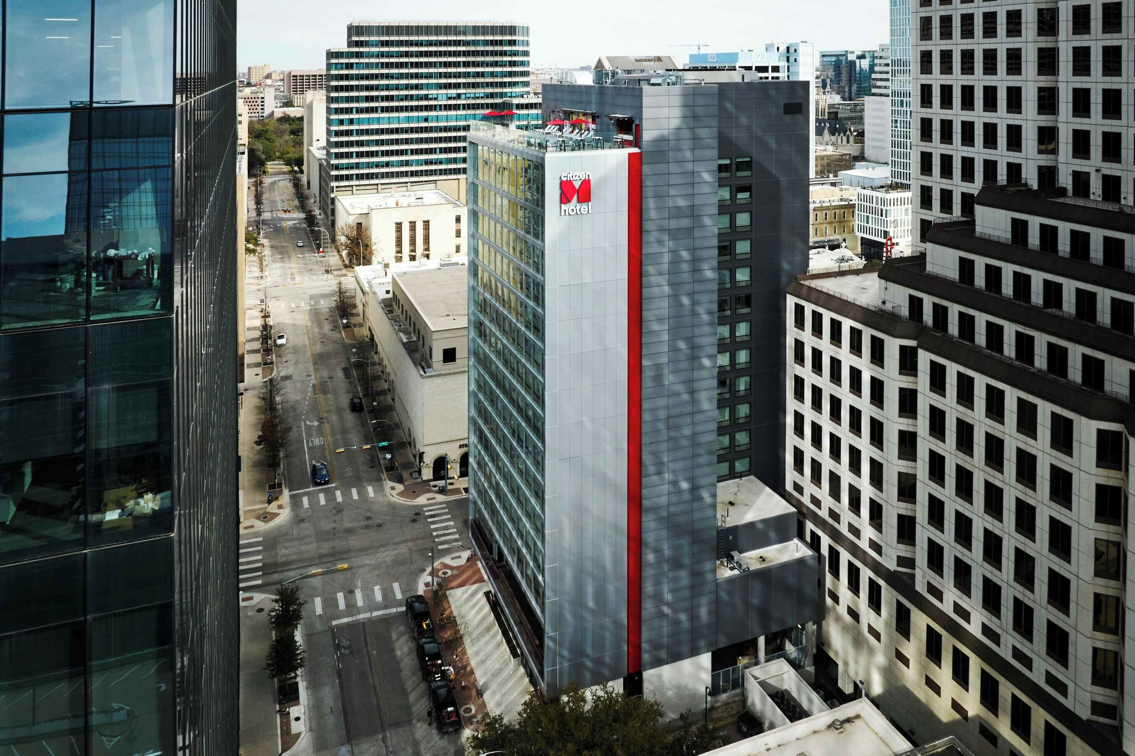 Un hotel modern amb un logo vermell i blanc destacat, envoltat d'edificis alts i una vista clara del carrer.