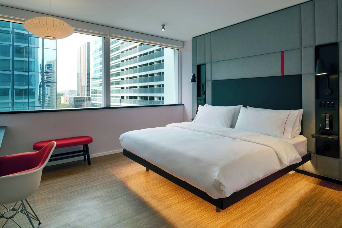 A modern hotel room with a large bed, a red chair, and a view of tall buildings through a large window.