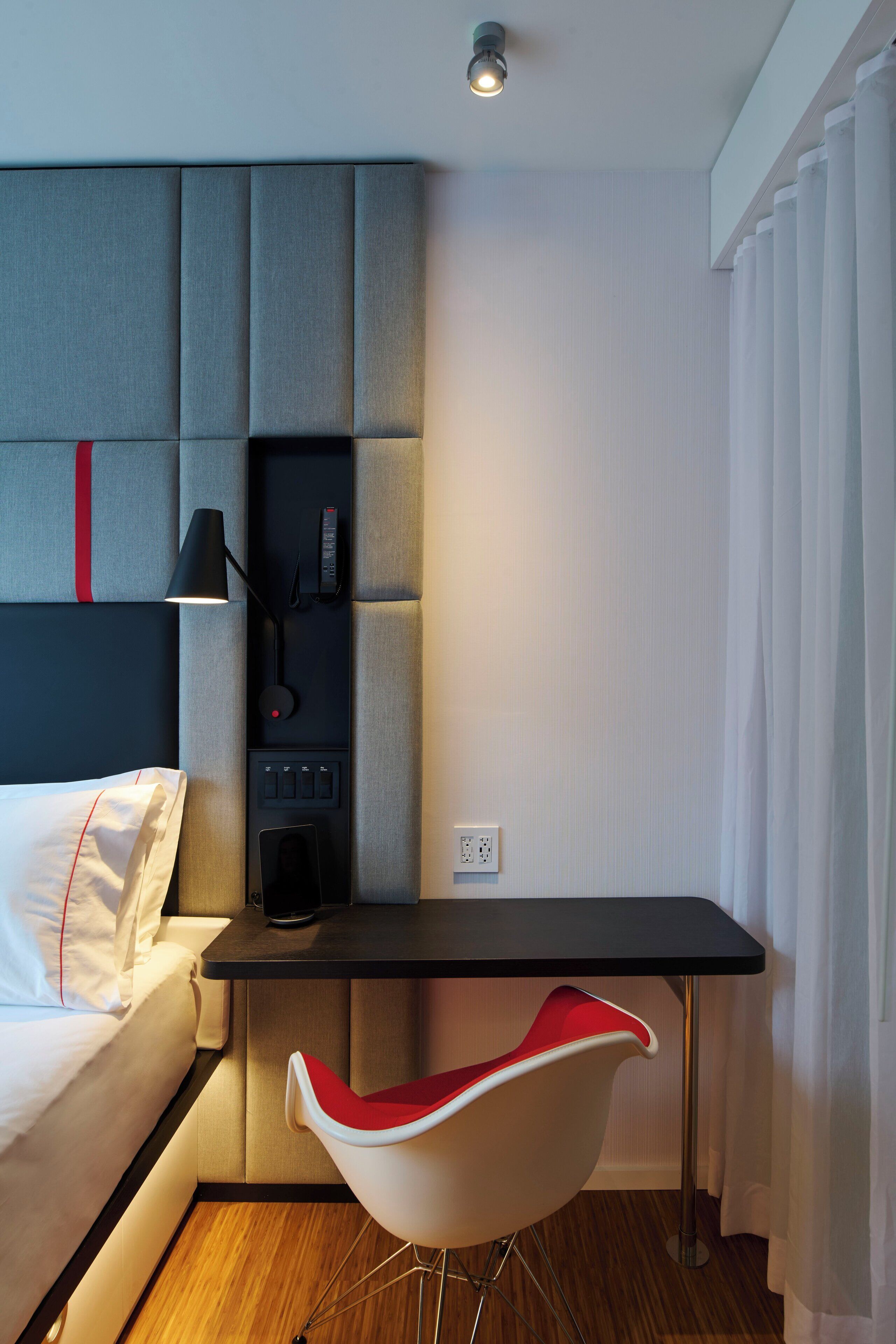 A modern hotel room with a bed, a desk, a chair, and a control panel.