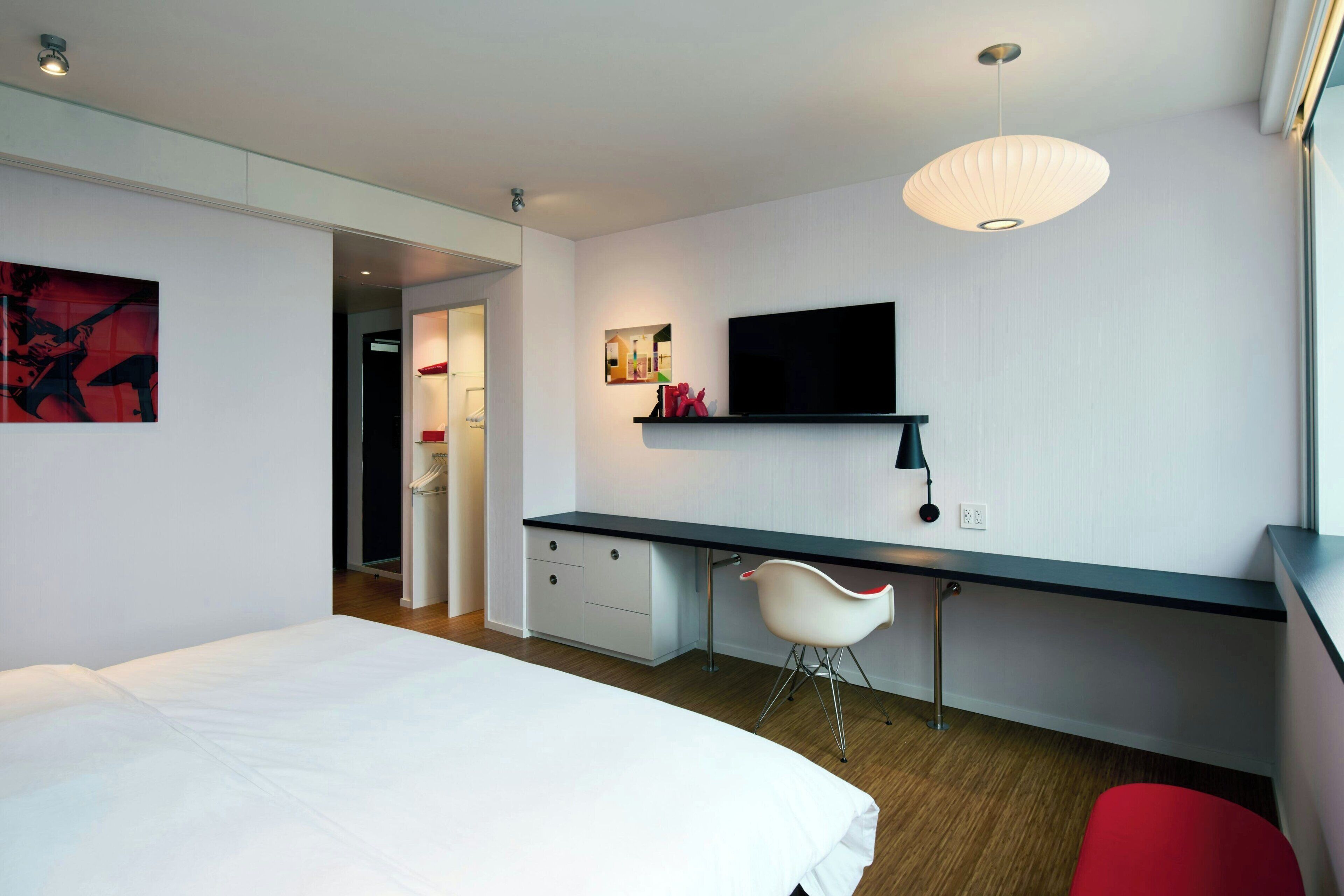 A modern hotel room with a bed, a desk, a chair, a TV, and a red accent chair.