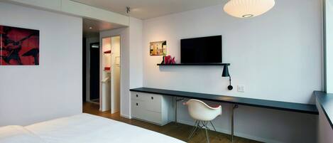A modern hotel room with a bed, a desk, a chair, a TV, and a red accent chair.